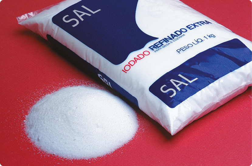 Fotografia. Um pacote de sal de cozinha, com sal despejado ao lado dela. Na embalagem está escrito: Sal. Iodado refinado extra. Para mesa e cozinha. Peso líquido 1 quilograma.