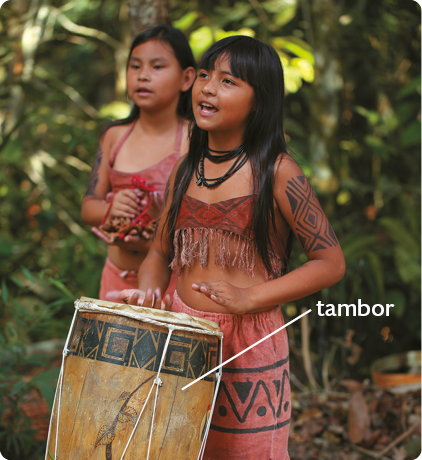 Fotografia de duas meninas com cabelos pretos e lisos, usando saias e blusas curtas, com grafismos indígenas nas roupas e nos braços; a menina à frente está usando as mãos para tocar um tambor, um instrumento cilíndrico de madeira.