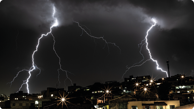 Fotografia de céu escuro com nuvens e casas abaixo. Entre o céu e as casas há dois raios ramificados.