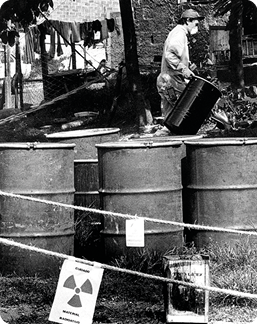 Fotografia em preto e branco de um local aberto, com árvores e uma casa ao fundo. Há uma pessoa com macacão e máscara segurando um tambor. À frente, tambores fechados e cordas isolando o local com placas com o símbolo da radioatividade.