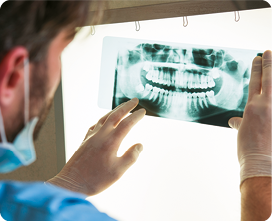 Fotografia de uma pessoa de costas, com luvas cirúrgicas, segurando uma radiografia de boca com as duas mãos sobre um quadro luminoso branco. Na radiografia há os contornos dos dentes, que aparecem com a cor branca.