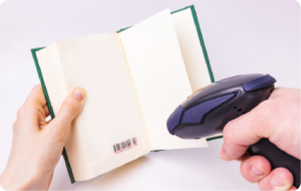 Fotografia. Duas mãos de uma pessoa. A mão esquerda está segurando um livro aberto com um código de barras em uma das folhas. A outra mão está segurando um objeto arredondado com cabo, apontando para o código de barras. Há uma linha vermelha sobre o código de barras.