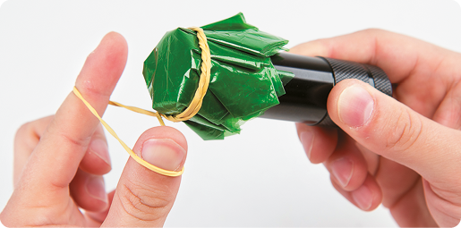 Fotografia. Duas mãos de uma pessoa. A mão direita está segurando uma lanterna cilíndrica com um papel-celofane verde na frende da lanterna. A outra mão está utilizando um elástico para prender o papel-celofane à lanterna.