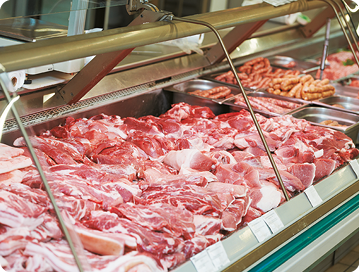 Fotografia. Uma vitrine de açougue com carnes expostas. Ela possui um formato retangular e painéis de vidro transparente. As carnes têm coloração vermelha e branca, dispostas em bandejas de metal. O local é bem iluminado, sendo possível observar frisos verdes em volta da vitrine.