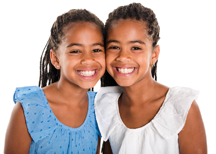 Fotografia de duas meninas sorrindo e olhando para frente, com cabelos pretos e longos com tranças, usando blusas sem mangas, uma azul e outra branca. Elas têm o formato do rosto, olhos, sobrancelhas, boca, nariz, orelhas e queixo muito parecidos.