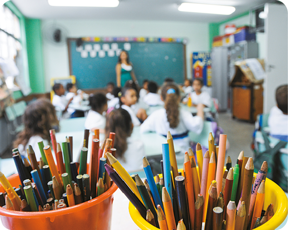 Fotografia 4. Destaque para potes repletos de lápis de cor em um sala de aula. Ao fundo, alunos sentados em carteiras escolares de frente para a professora e para a lousa.