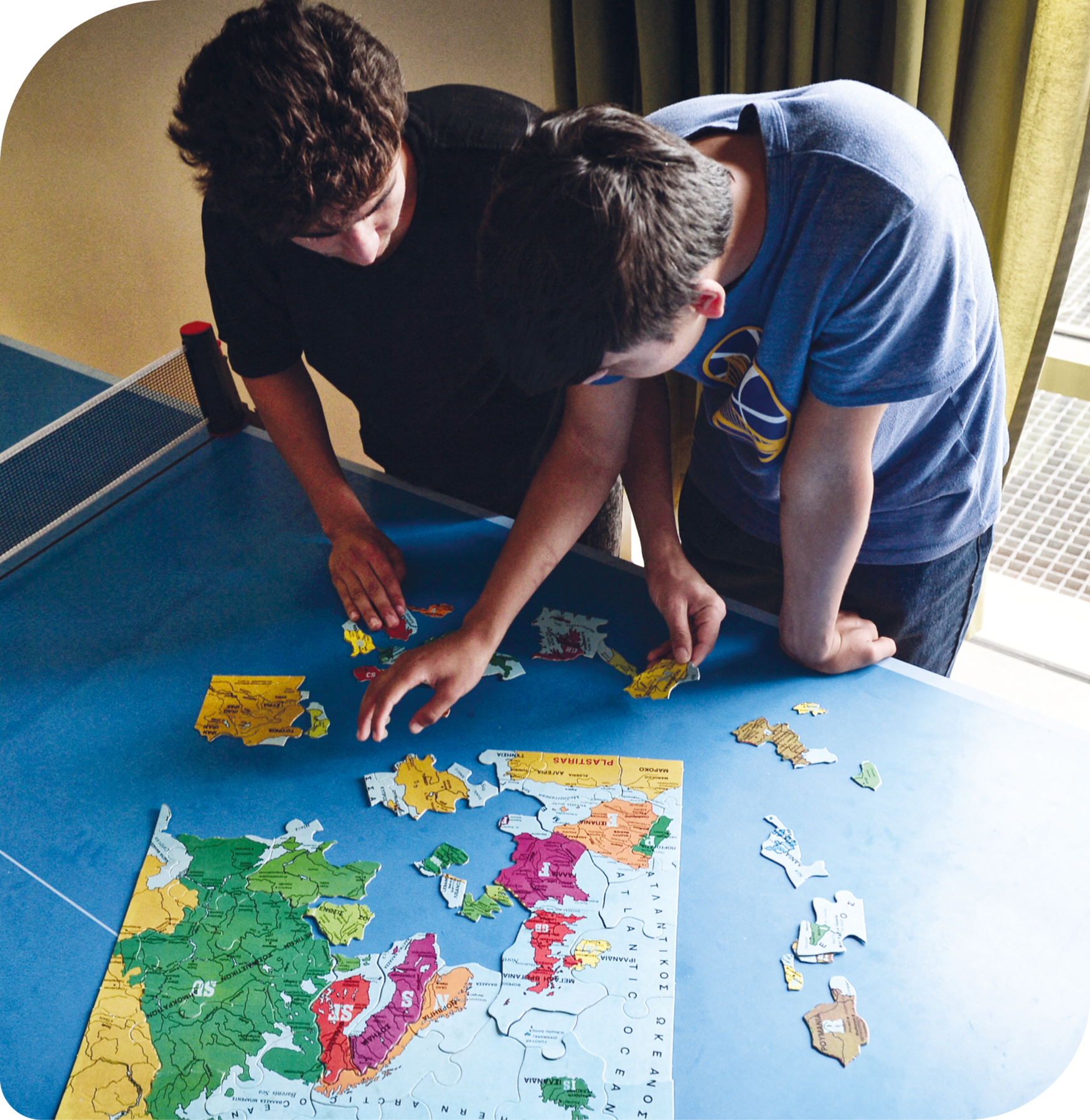 Fotografia. Dois rapazes em pé montando um quebra-cabeças com a figura de um mapa. Eles posicionam grupos de peças sobre uma mesa de pingue-pongue.