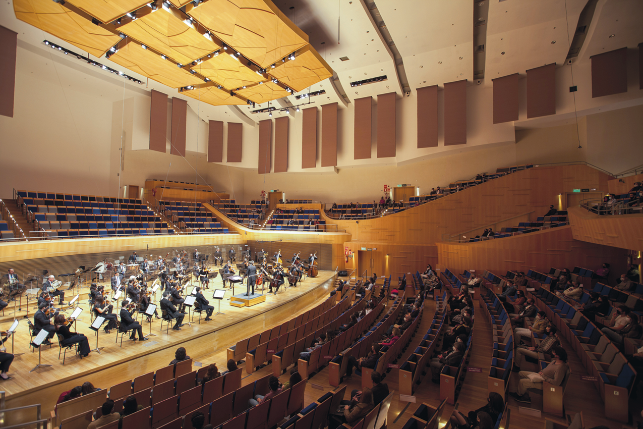 Fotografia. Uma orquestra em um palco cercado por fileiras de cadeiras com pessoas assistindo a apresentação.