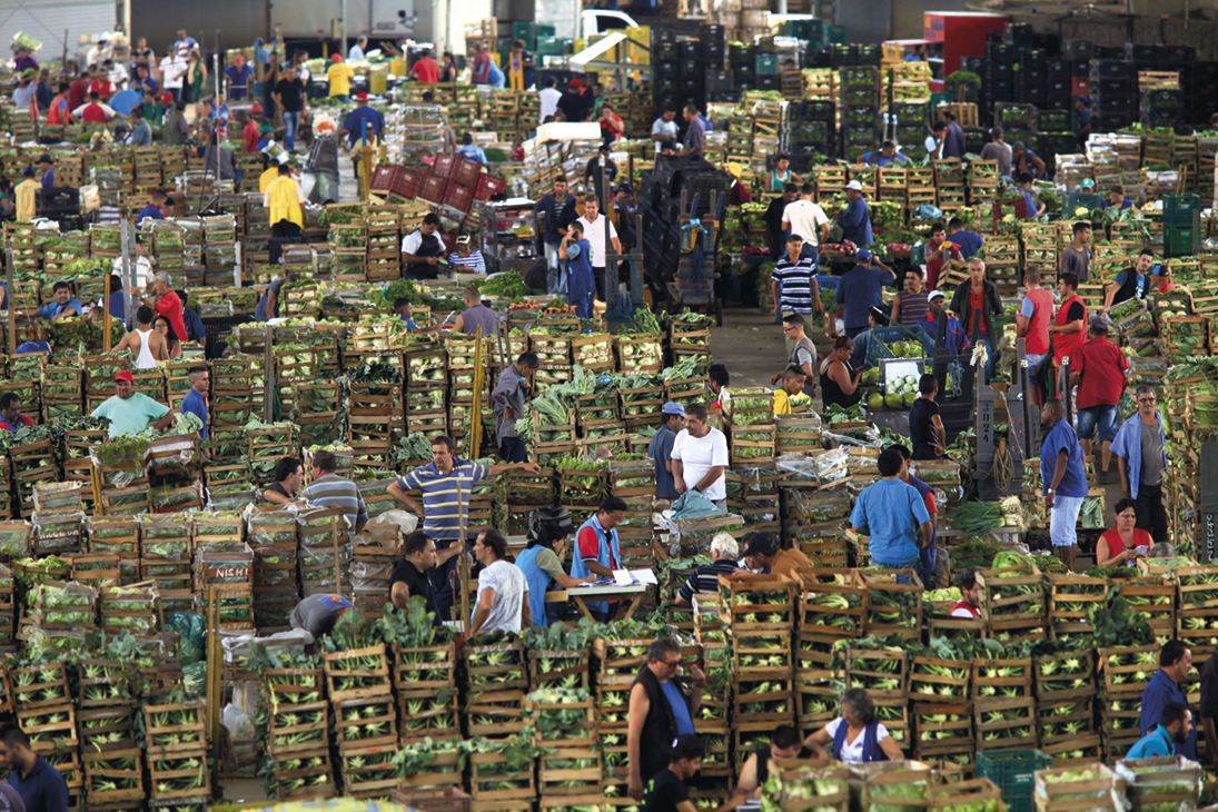 Fotografia. Um armazém repleto de caixas com verduras e muitas pessoas caminhando entre elas.