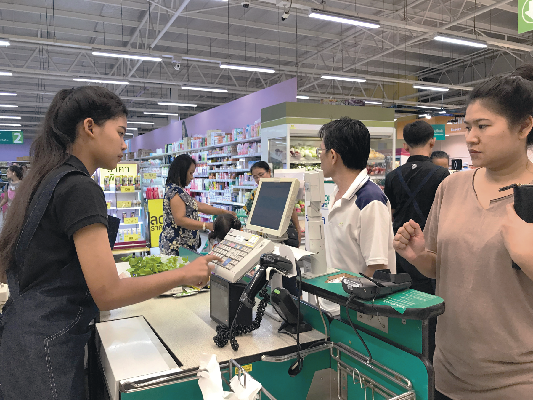 Fotografia B. Uma mulher em um caixa de supermercado digitando em teclas. Há uma fila de pessoas em frente a ela e ao fundo, pessoas entre prateleiras com produtos.