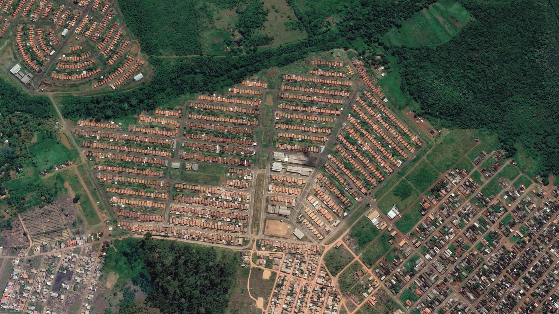 Fotografia. Imagem de satélite com diversas casas e construções dispostas em quarteirões ocupando a área central, sudeste e noroeste. Ao redor há áreas cobertas por vegetação.