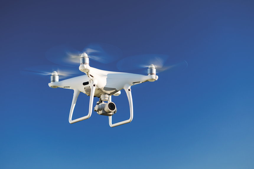 Fotografia. Um drone com uma câmera posicionada na parte inferior e três estruturas com hélices na parte superior.