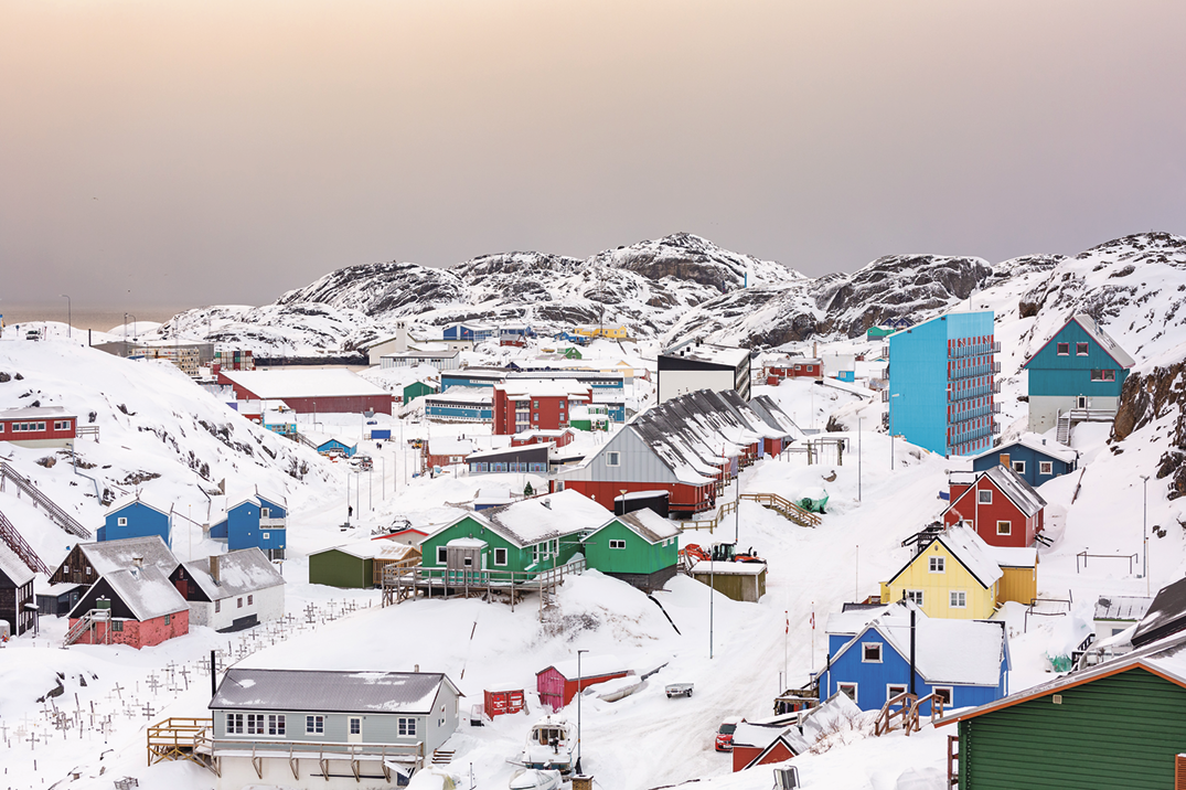 Fotografia. Uma cidade com pequenas casas coloridas parcialmente cobertas por neve. Ao fundo, montanhas rochosas também cobertas por neve.