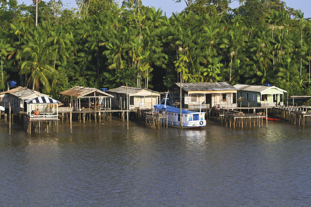 Fotografia. Casas de palafita sobre um rio largo. Ao fundo há árvores. Há um barco ancorado ao lado das casas.