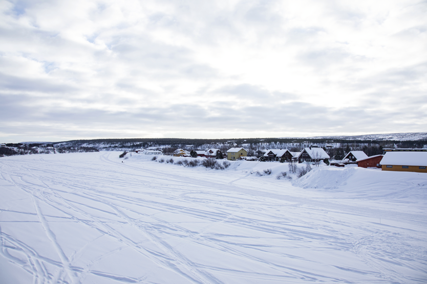 Fotografia A. Uma via coberta por neve. Ao fundo há algumas casas com os telhados também cobertos por neve e árvores.