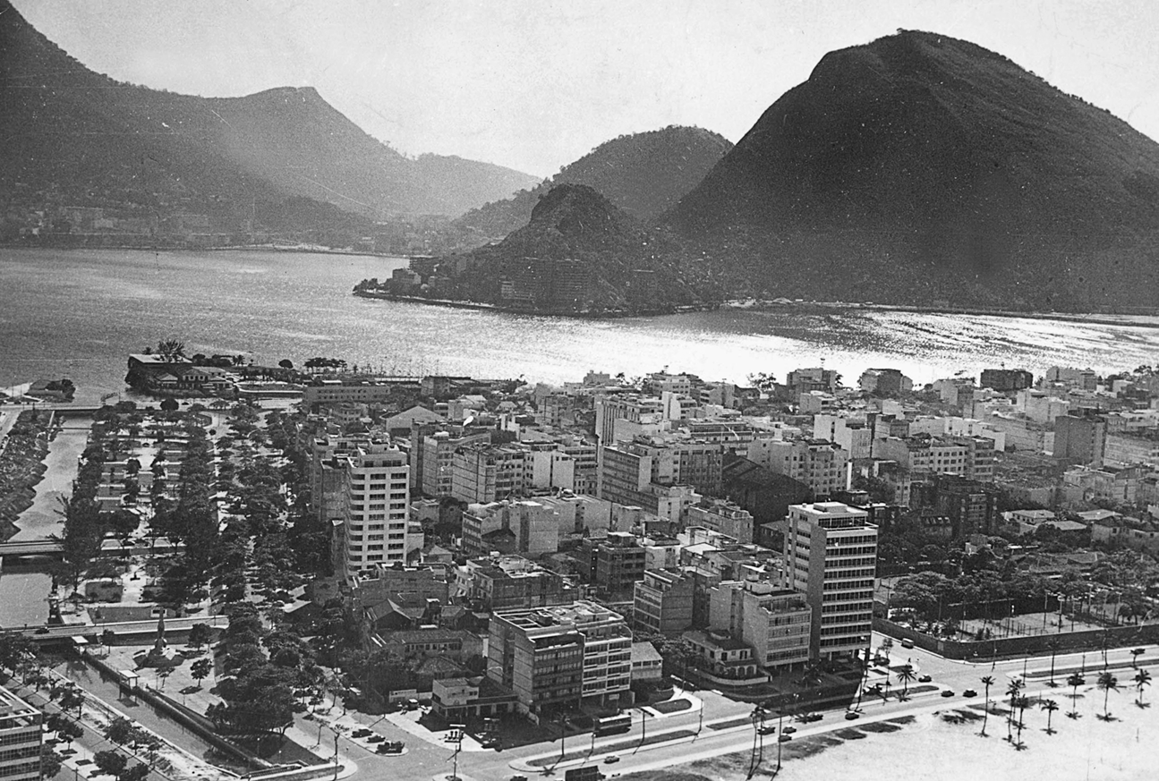 Fotografia em preto e branco. Vista de cima. No primeiro plano, cidade com prédios na orla de uma praia. Ao fundo, morros rochosos e o mar.