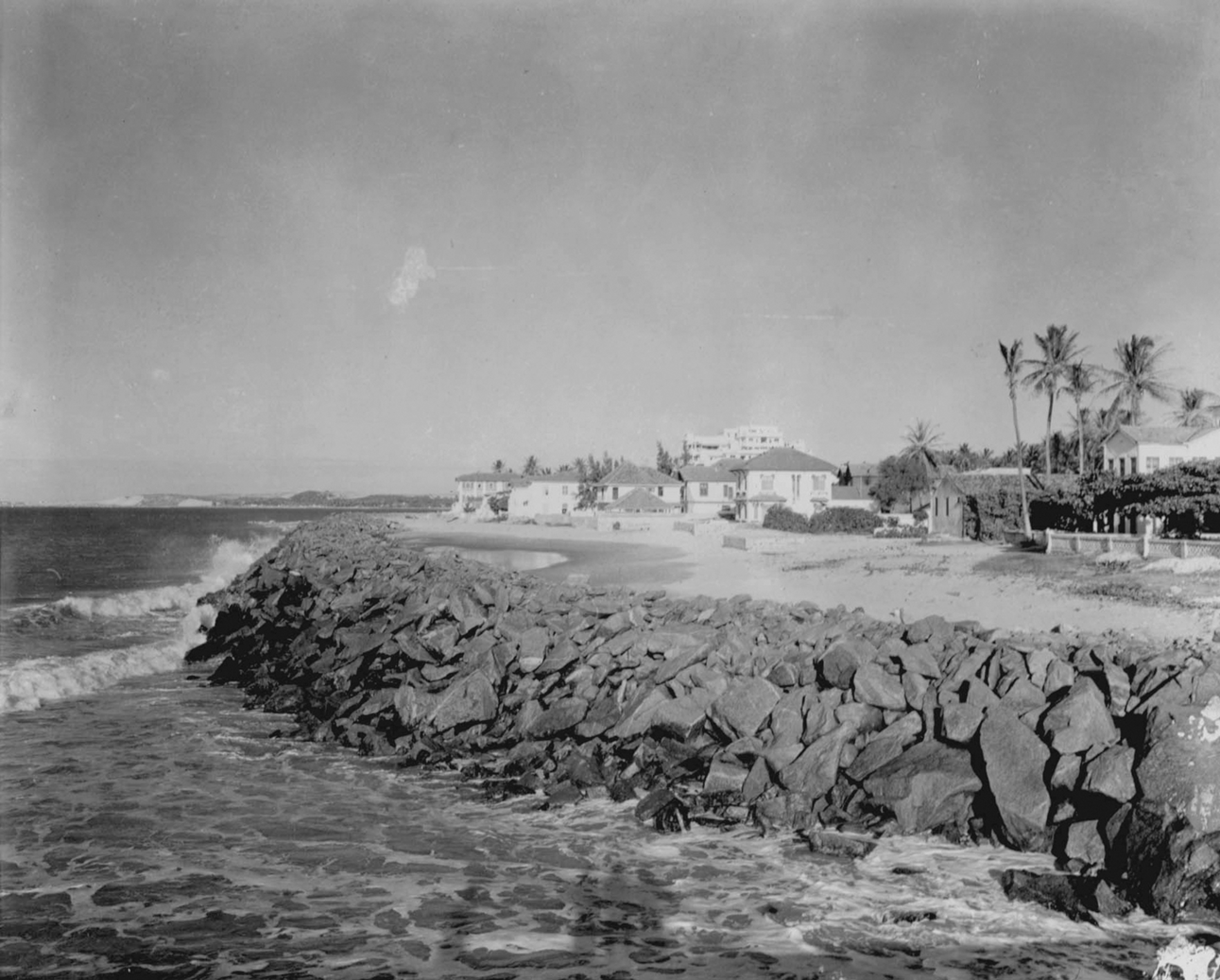Fotografia em preto e branco. Praia com a orla repleta de rochas. Acima delas há uma via, casas e palmeiras.