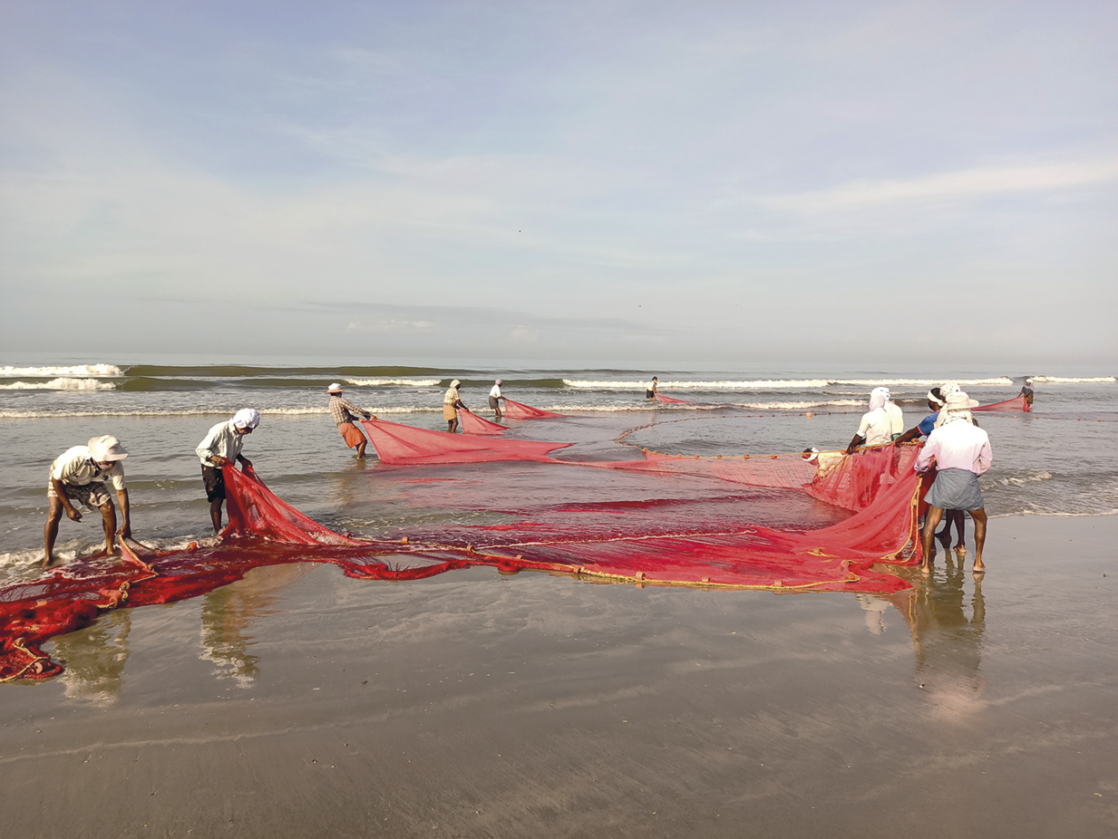 Fotografia. Pescadores esticando uma rede de pesca vermelha em uma praia. Ao fundo, o mar.