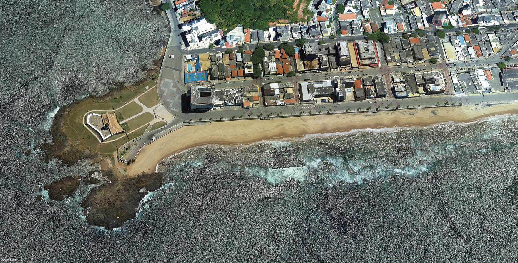 Fotografia. Vista aérea. À esquerda, um farol entre rochas em uma península. Ao fundo, construções em quarteirões em frente ao mar.