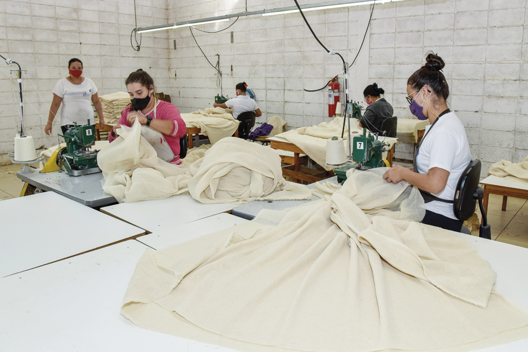 Fotografia. Mulheres trabalhando sentadas em frente a máquinas de costura. Há tecidos entre elas.