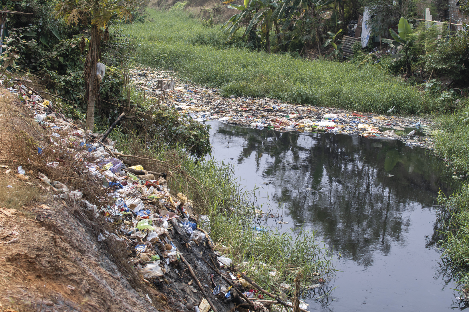 Fotografia. Curso de um rio repleto de lixo nas duas margens. Ao fundo, vegetação rasteira e árvores.