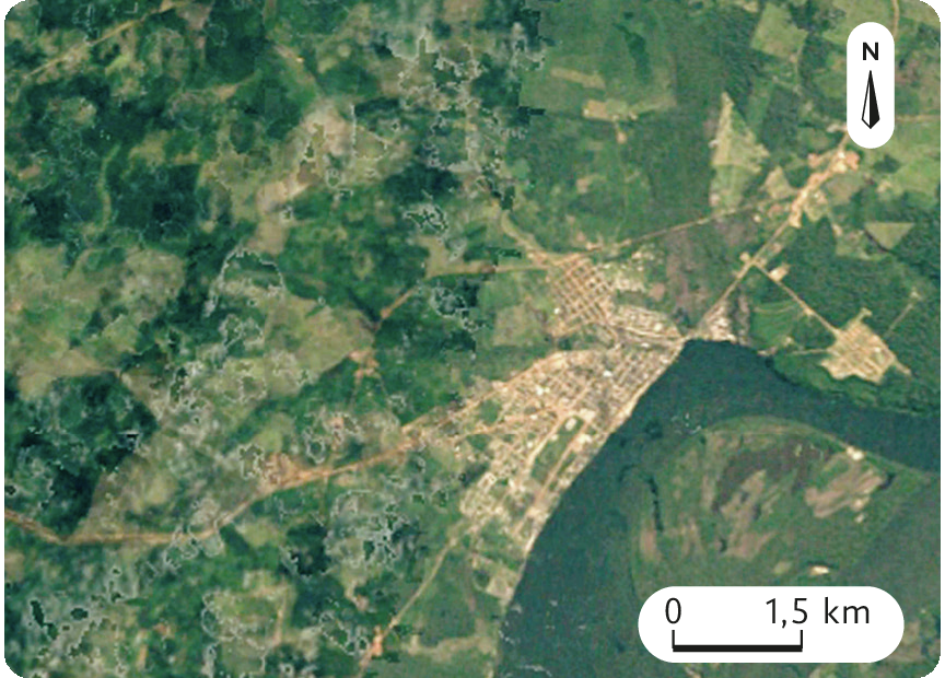 Fotografia. Vista aérea.  Imagem de satélite de uma cidade em meio a áreas de plantio e florestas. No canto superior direito, uma seta aponta para o norte e abaixo, a escala: 1,5 quilômetro por centímetro.