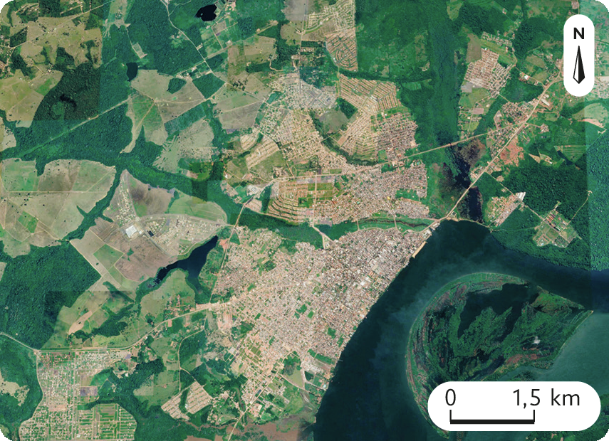 Fotografia. Imagem de satélite de uma grande cidade em meio a áreas de plantio e florestas. No canto superior direito, uma seta aponta para o norte e abaixo, a escala: 1,5 quilômetro por centímetro.
