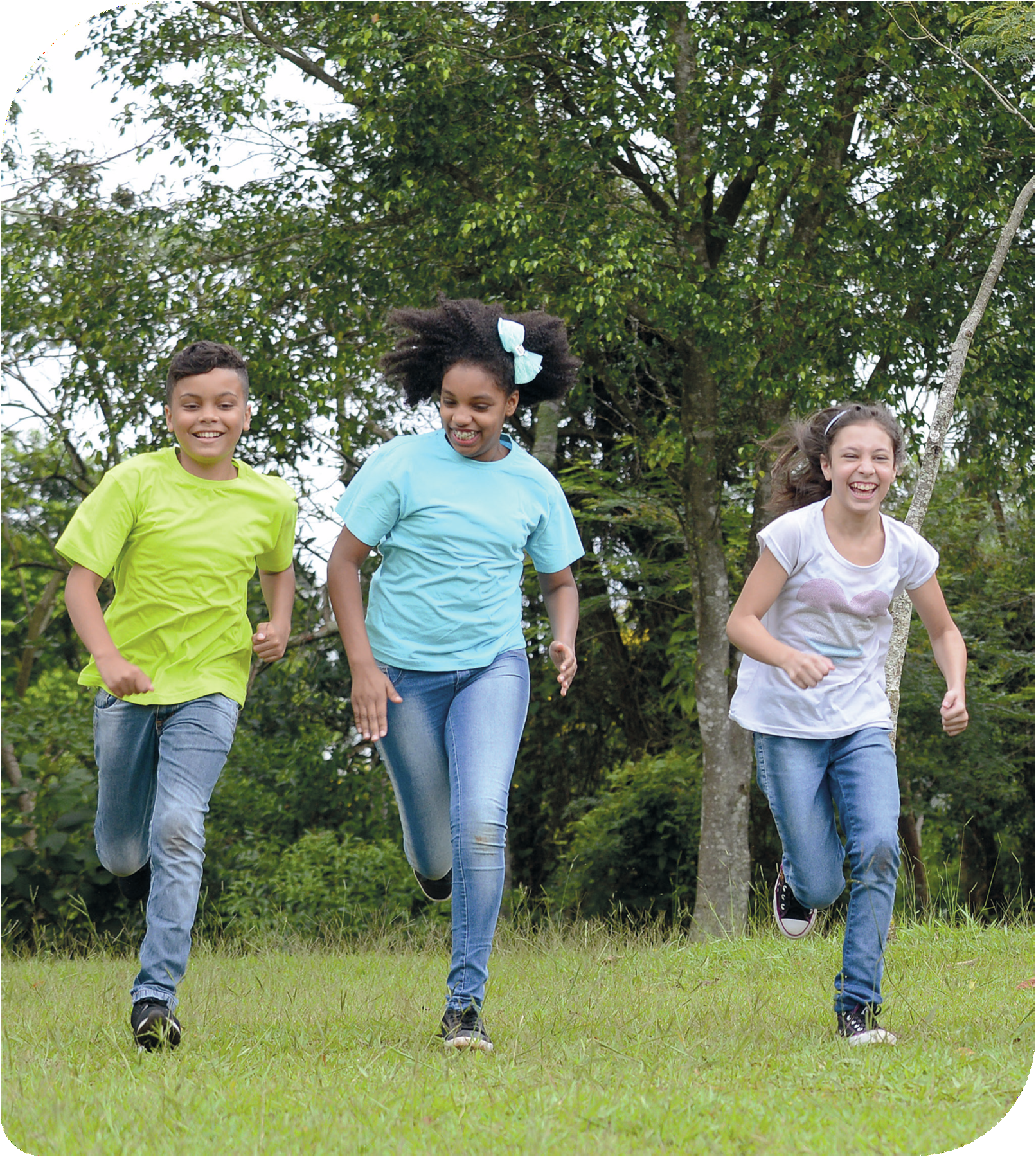 Fotografia. Três crianças correndo em uma área gramada. Elas sorriem. Ao fundo há árvores.