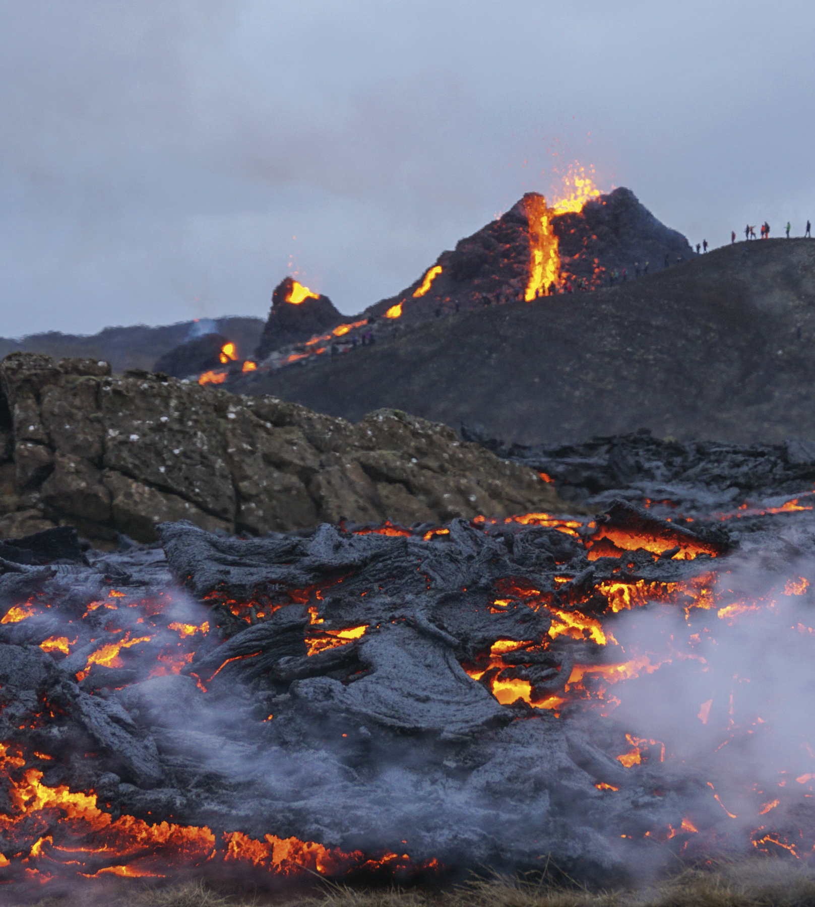 Fotografia. Destaque para um vulcão em erupção. Há lava sendo expelida e no solo onde está esfriando, em uma coloração acinzentada.