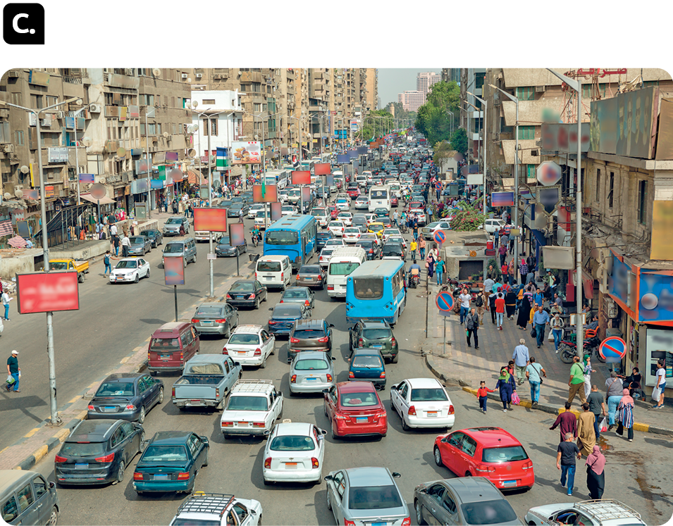 Fotografia C. Rua larga repleta de carros, com muitas pessoas nas calçadas. Há construções amplas nas laterais.