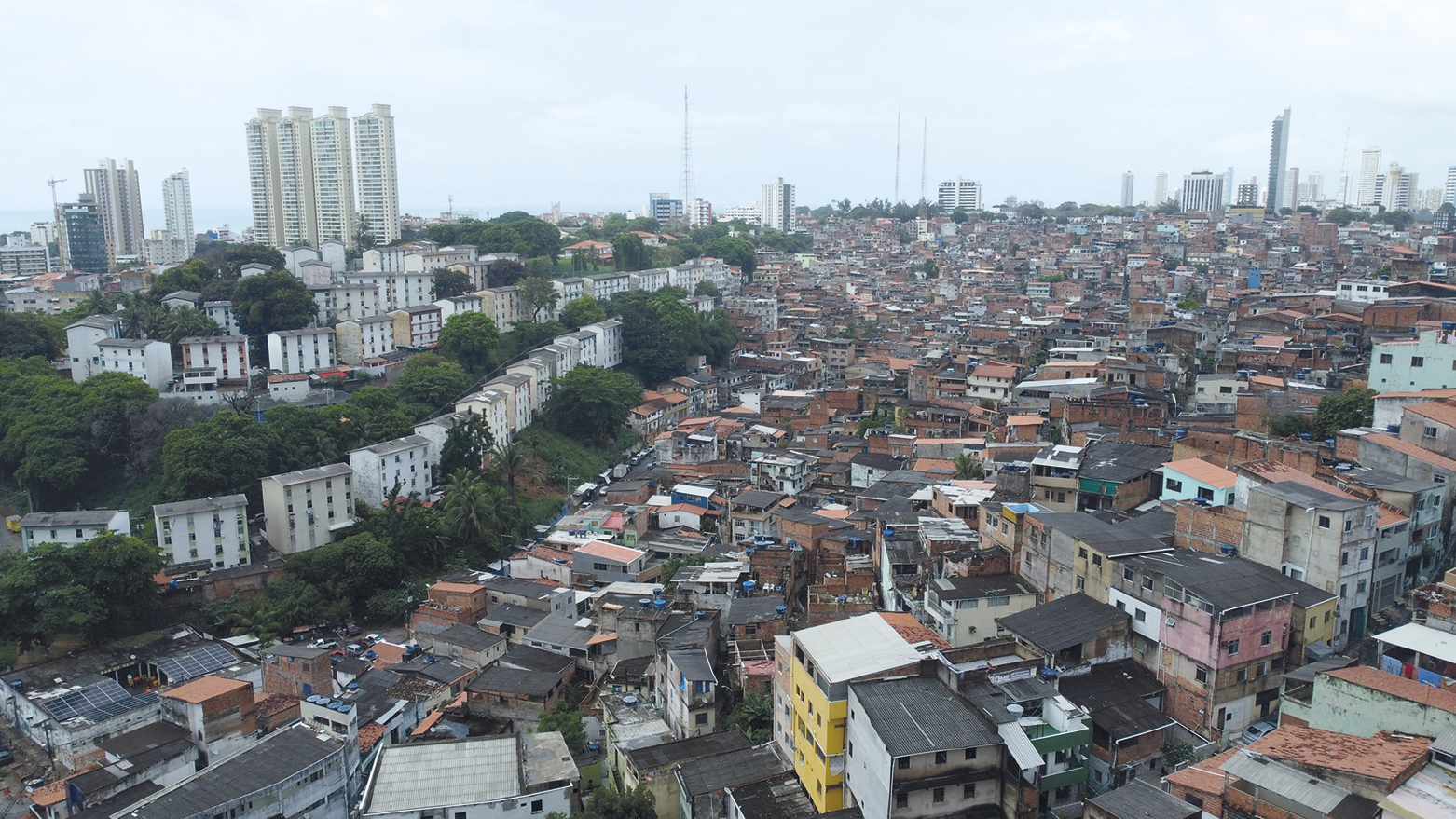 Fotografia. Uma cidade vista de cima com casas pequenas amontoadas à direita, e com prédios entre árvores à esquerda.