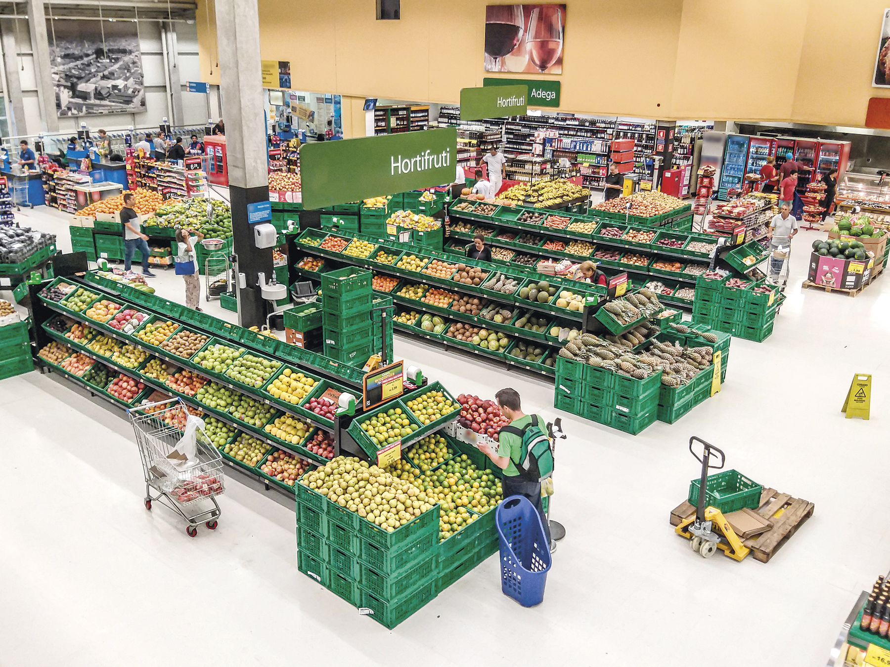 Fotografia. Vista aérea de um supermercado com frutas e legumes expostos em caixas verdes em uma área sinalizada como: HORTIFRÚTI. Ao redor há mais prateleiras.