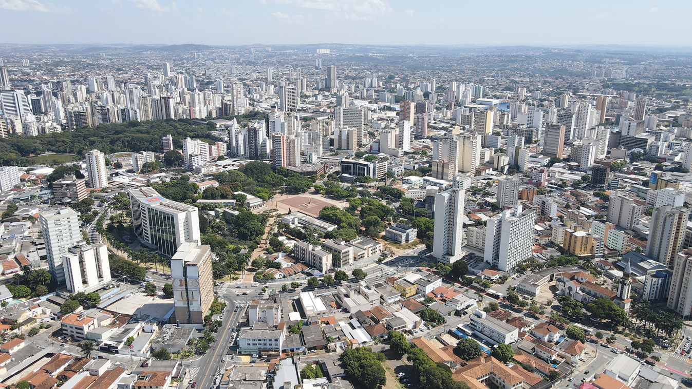 Fotografia. Vista aérea de uma cidade repleta de prédios e casas, com uma praça central. Há árvores dispersas entre as construções e as ruas.