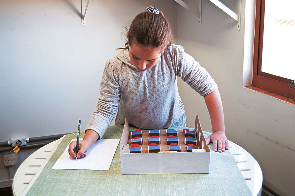 Fotografia. Uma menina em pé ao lado de uma maquete. Ela está fazendo anotações em uma folha de papel.