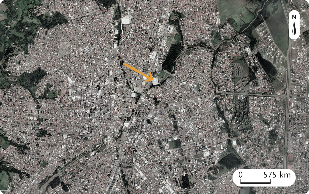 Fotografia B. Imagem de satélite. A cidade vista de uma distância maior com a seta apontando para terminal rodoviário ao centro. No canto superior, seta apontando para o norte. Abaixo, a escala: 575 quilômetros por centímetro.