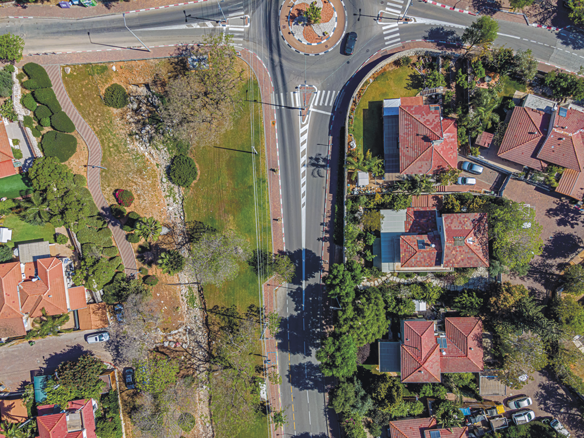 Fotografia. Vista aérea. Uma rotatória com uma área gramada à esquerda, e casas à direita, com a via passando ao centro.