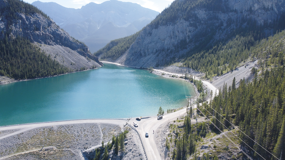 Fotografia. Um lago cercado por montanhas e pinheiros. Há uma via com carros à esquerda.