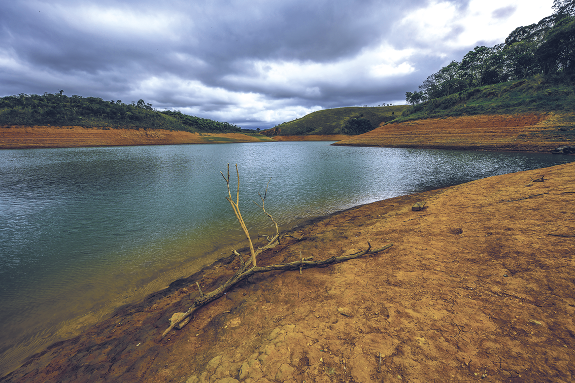 Fotografia. Margem de uma represa com terra seca e árvores ao fundo.