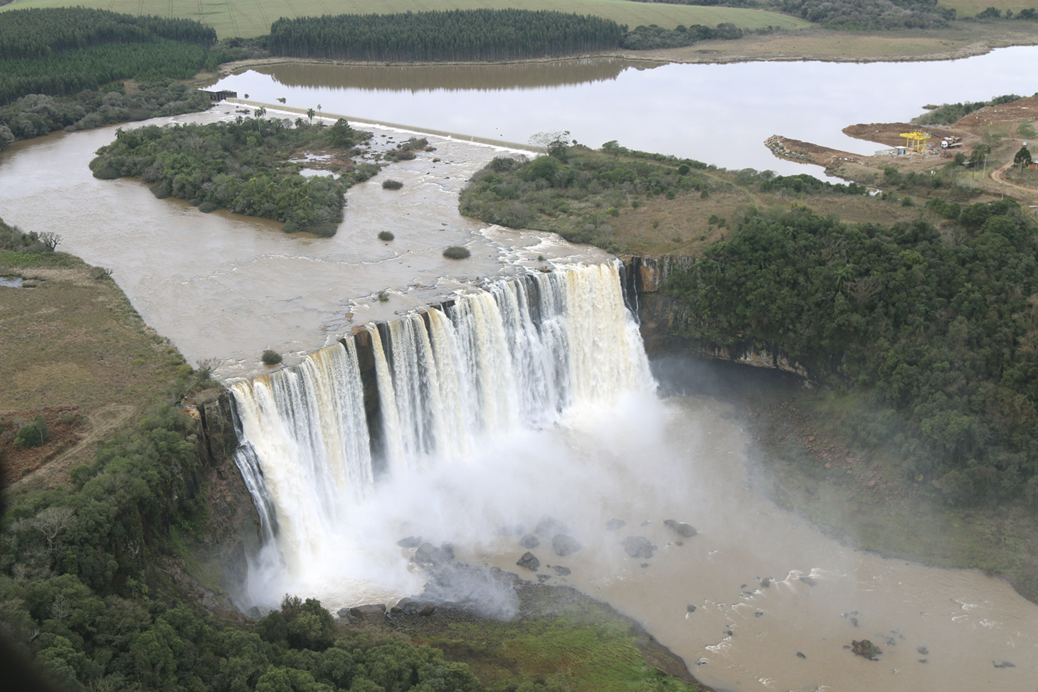 Fotografia. Vista de cima. Um rio desaguando e uma grande cachoeira. Ao redor há mata.