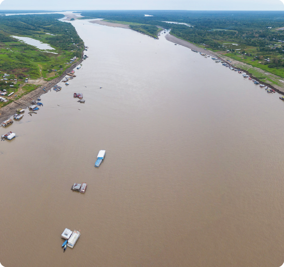 Fotografia. Um extenso rio com algumas embarcações. Há vegetação nas margens.