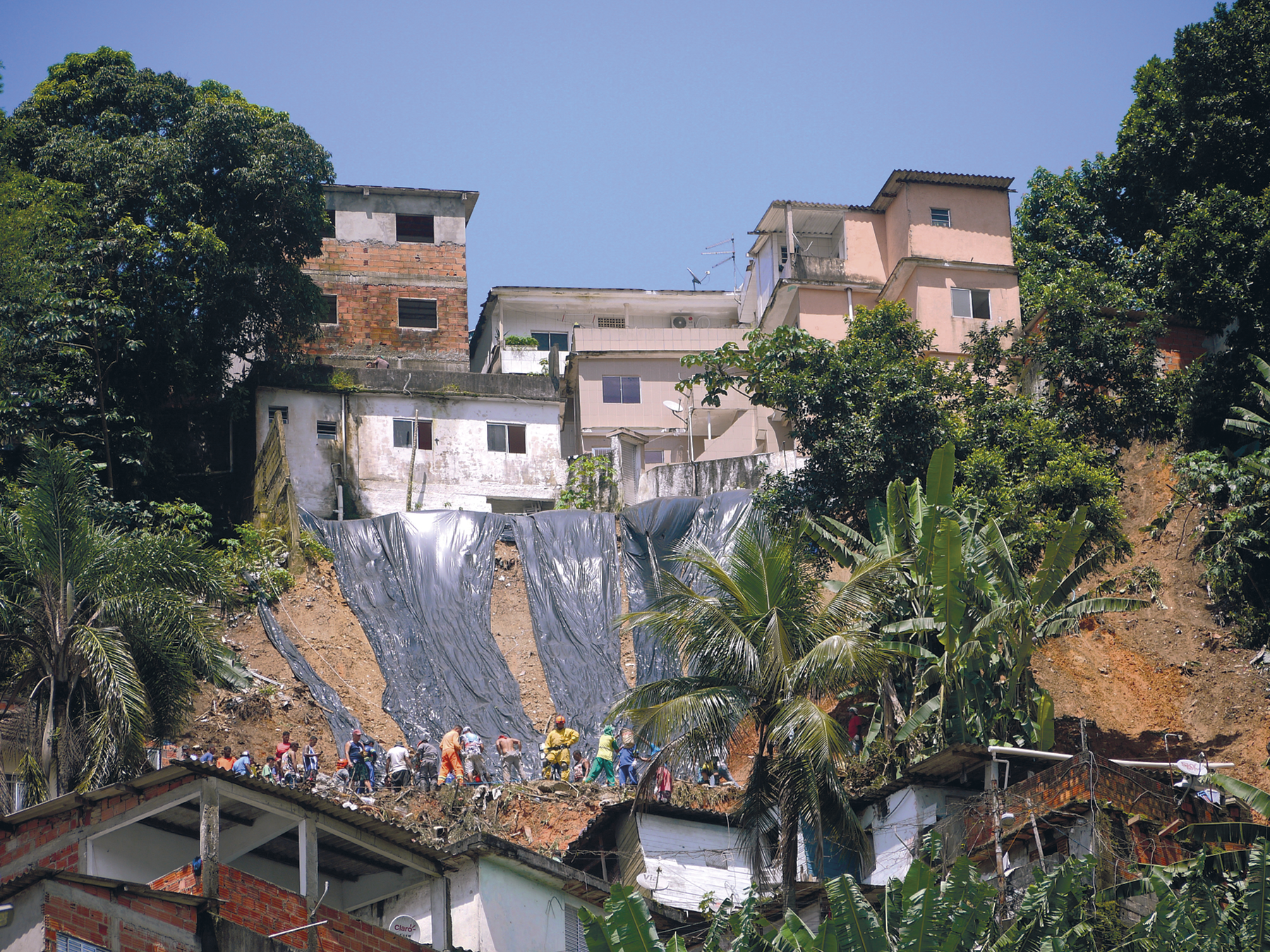 Fotografia. Deslizamento de terra na encosta de um morro. Há casas no topo e abaixo, pessoas reunidas. Sobre o morro há lonas e, nas laterais há árvores.