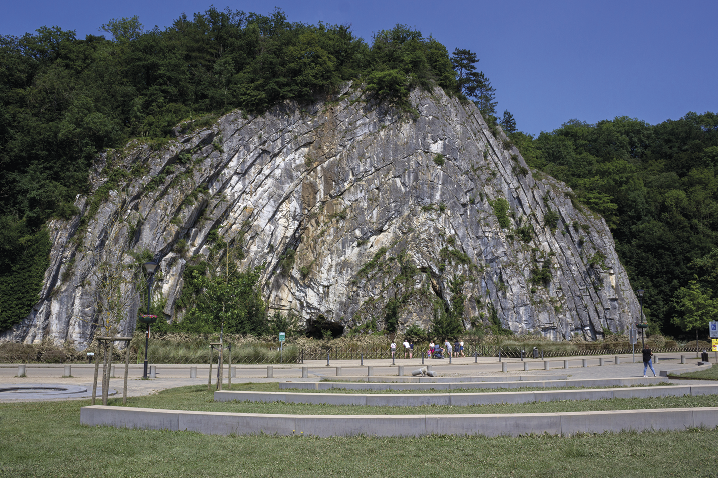 Fotografia. Uma grande rocha com ranhuras e camadas em formato arqueado, com árvores no topo e algumas pessoas a frente.