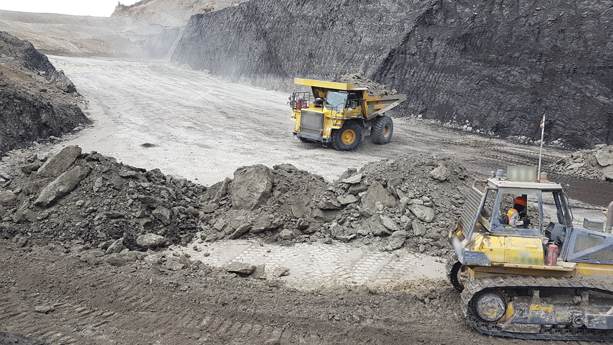 Fotografia. Tratores em uma mina com resíduos e rochas de cor cinza.
