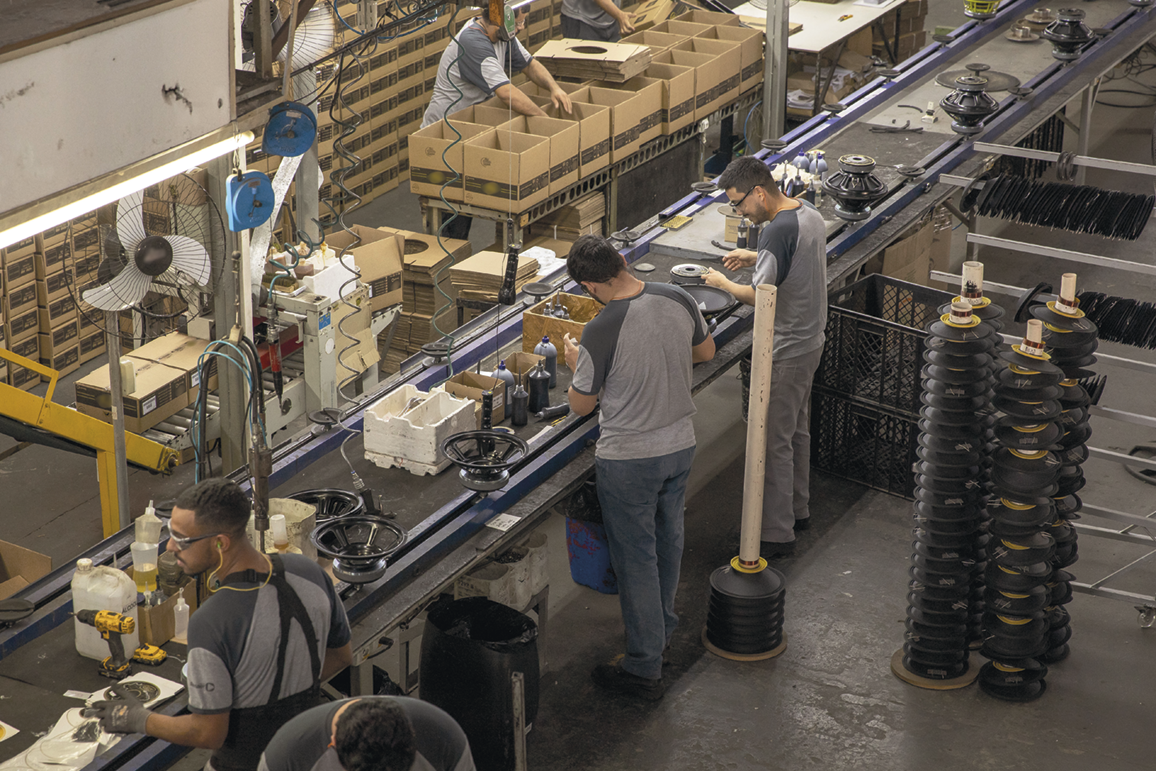 Fotografia. Pessoas trabalhando em uma linha de produção. Eles estão em pé em frente a uma esteira com produtos. Ao fundo há mais pessoas manuseando caixas.