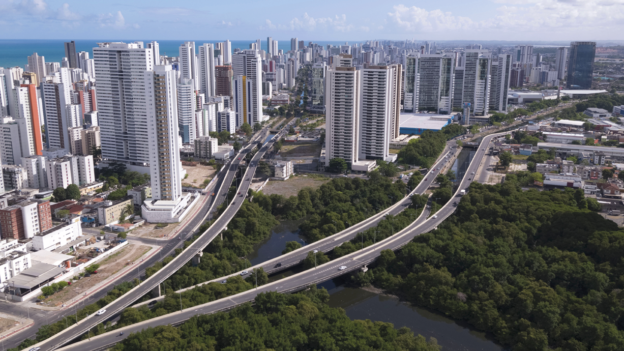 Fotografia. Vista de cima. Vias entre vegetação e rios que passam por uma cidade com muitos edifícios.