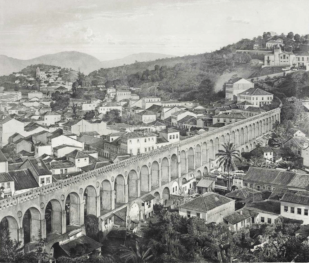 Fotografia em preto e branco. Vista de cima. Muitas casas amontoadas. Entre elas há dutos em duas colunas de arcos sobrepostas. Ao fundo, morro com mais casas.