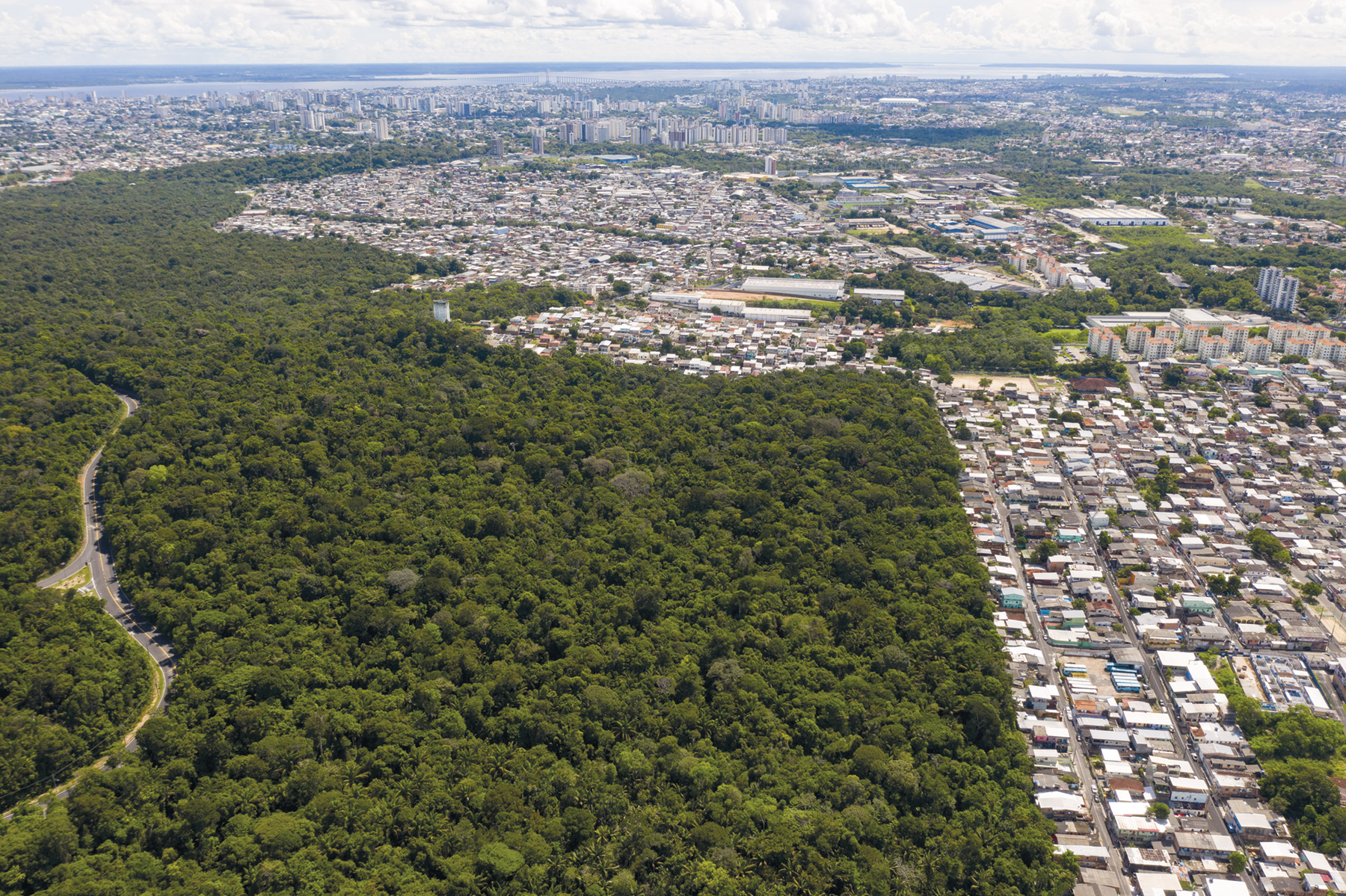 Fotografia. Vista de cima. À esquerda, floresta densa. À direita e ao fundo, cidade com muitas construções.