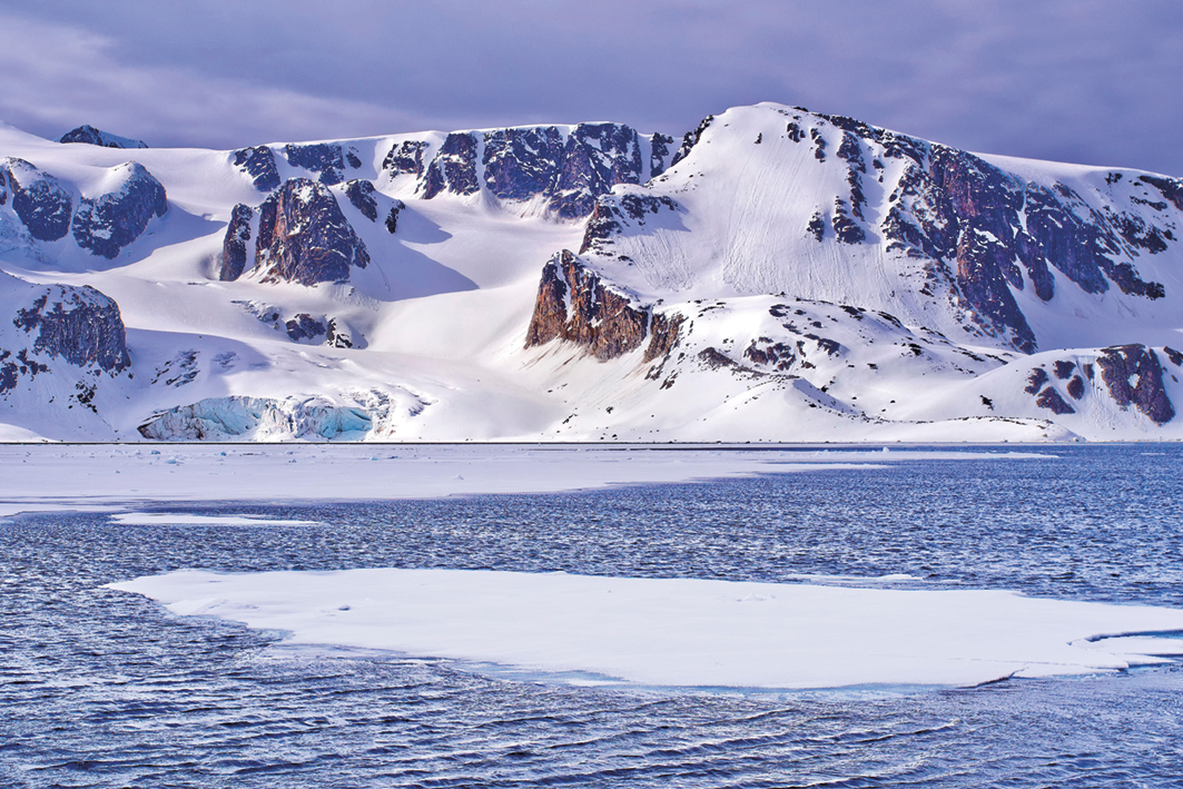 Fotografia. No primeiro plano, o mar. Ao fundo, formações rochosas cobertas por neve.