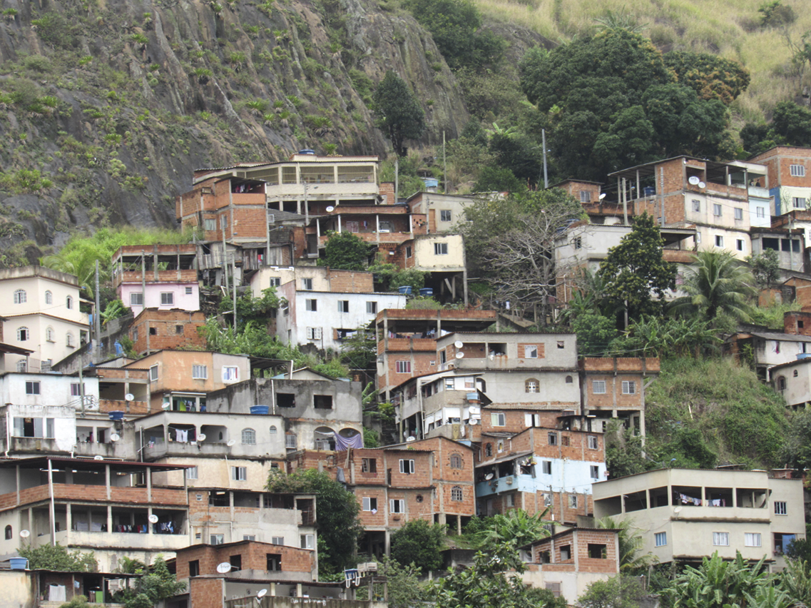 Fotografia. Casas amontoadas na encosta de um morro coberto por vegetação. Há algumas árvores entre as casas.