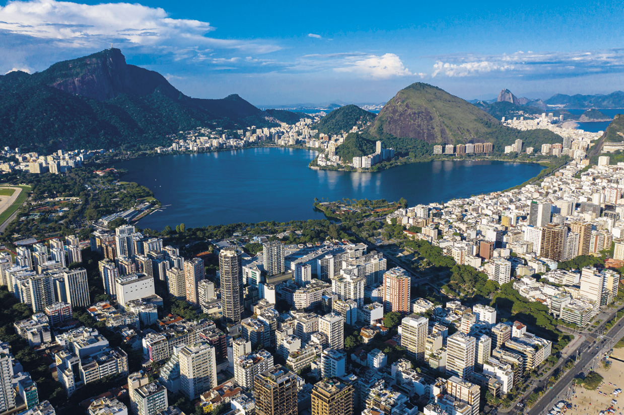 Fotografia. Vista de cima. Cidade com muitos edifícios ao redor de uma baía. Ao fundo há morros rochosos.